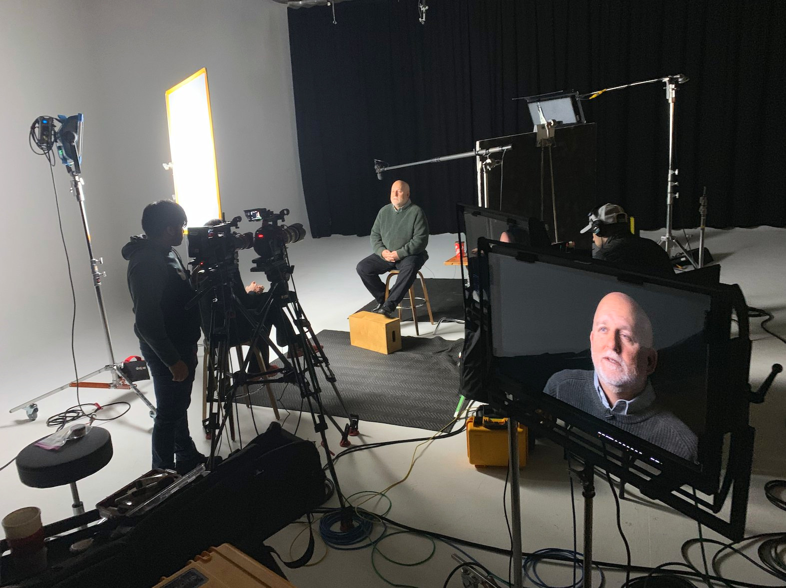 Two cameras, lighting, a boom microphone, and a monitor around a seated interview subject