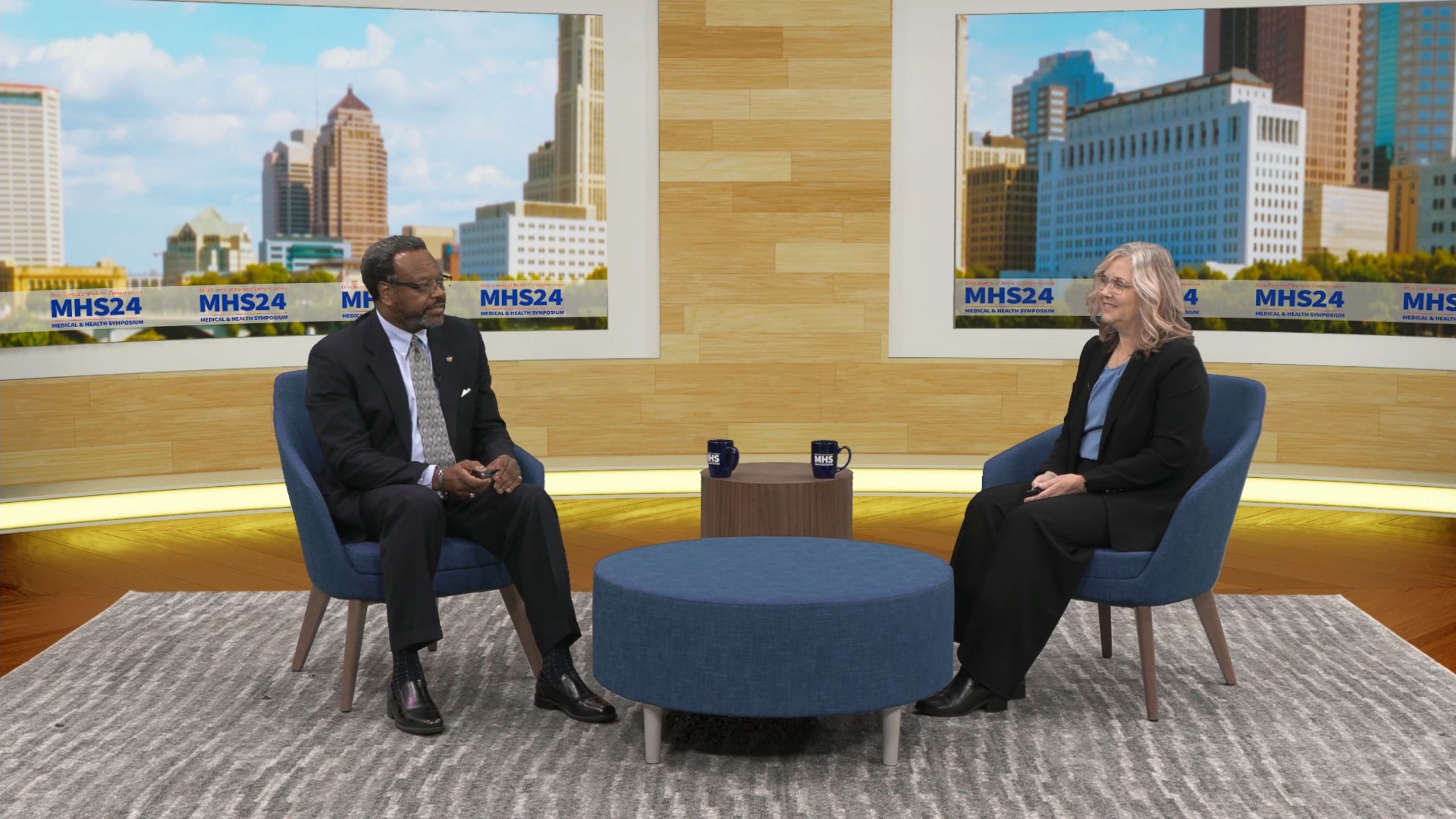 Two presenters in Ohio BWC’s finished virtual studio, with symposium branding and a Columbus skyline backdrop